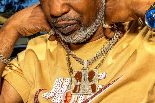 Close-up of an African American man wearing gold jewelry, including a necklace with a large pendant, and a gold watch.