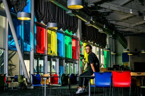 Adult male sitting on a colorful chair indoors with natural light streaming through large windows with red, yellow, green, and blue glass.