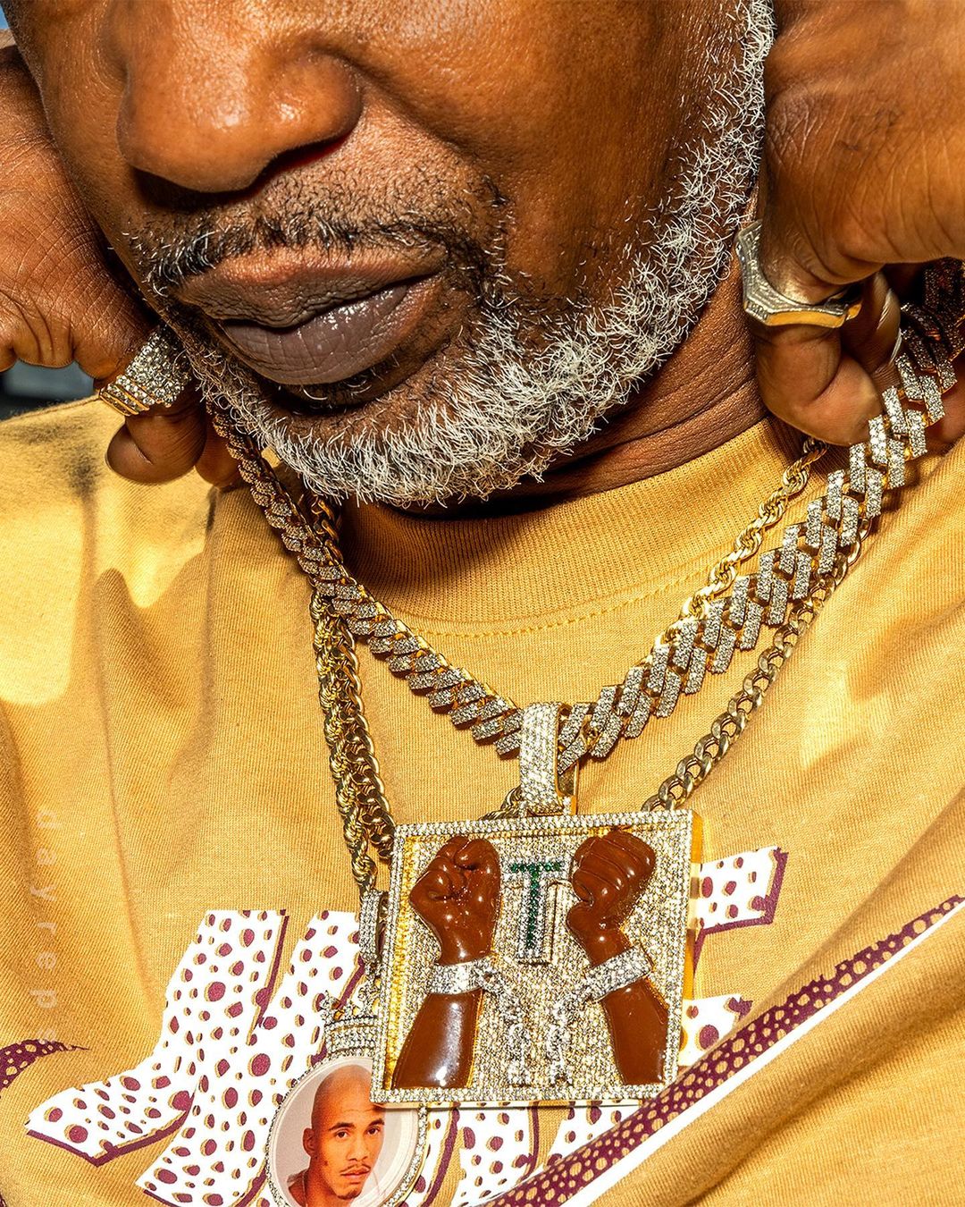 Close-up of a man with gold chains and a pendant featuring two fists, wearing a patterned shirt. He has short gray hair and a goatee.