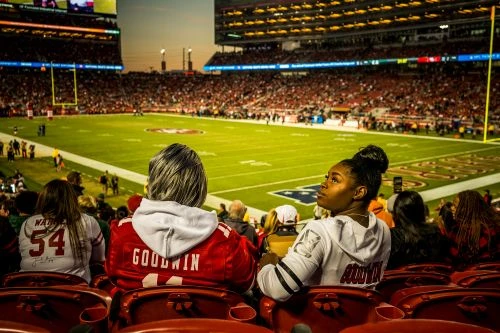 Fans in stadium seats watching American football game; spectators wear team jerseys, field is green and filled with players.