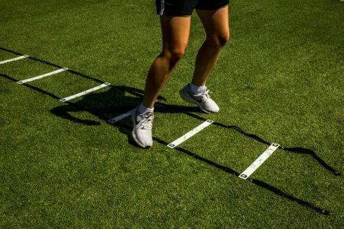 Athlete's legs and feet run through a speed and agility ladder on artificial grass in bright daylight, enhancing sports training.