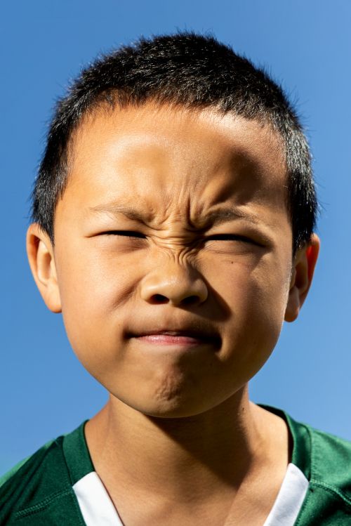 Close-up portrait of an Asian child with a scrunched, concentrated face against a bright blue sky, wearing a green and white sports jersey.