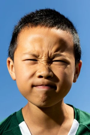 Close-up portrait of an Asian child with a scrunched, concentrated face against a bright blue sky, wearing a green and white sports jersey.