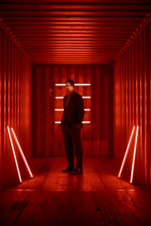 Man in a suit stands in a long corridor with red walls and floor, lit by neon lights forming a rectangle and lines.
