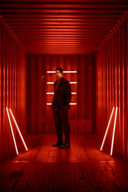 Man in a suit stands in a long corridor with red walls and floor, lit by neon lights forming a rectangle and lines.
