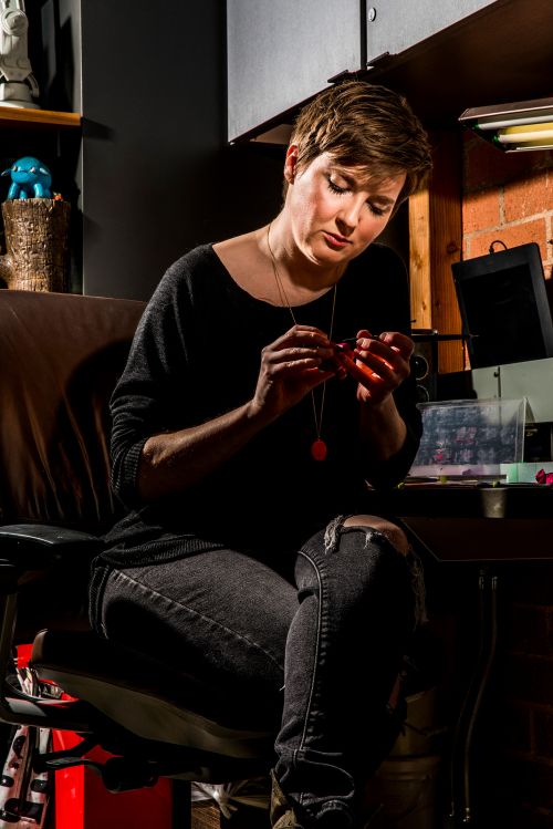 Adult female working on jewelry design at a desk in a studio with a brick wall, wearing black top and distressed jeans with soft lighting.