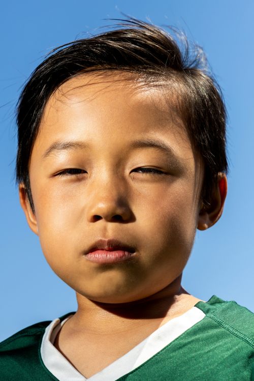 Close-up portrait of an Asian child with dark hair, wearing a green athletic jersey, set against a clear, bright blue sky.