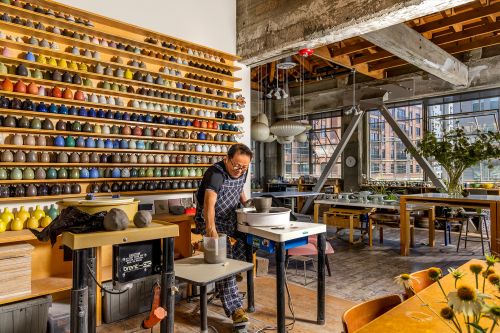 Adult male potter at his wheel, in a sunny, spacious studio surrounded by rows of colorful ceramics and pottery equipment, indoor setting.
