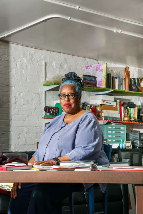 Portrait of a mature Black woman seated at a desk in an indoor room with shelves behind her, wearing glasses and a blue top.
