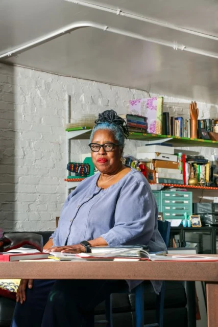 Portrait of a mature Black woman seated at a desk in an indoor room with shelves behind her, wearing glasses and a blue top.