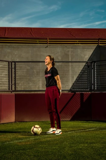 A woman in athletic wear stands laughing on a soccer field with a soccer ball at her feet, photographed outdoors on a sunny day.