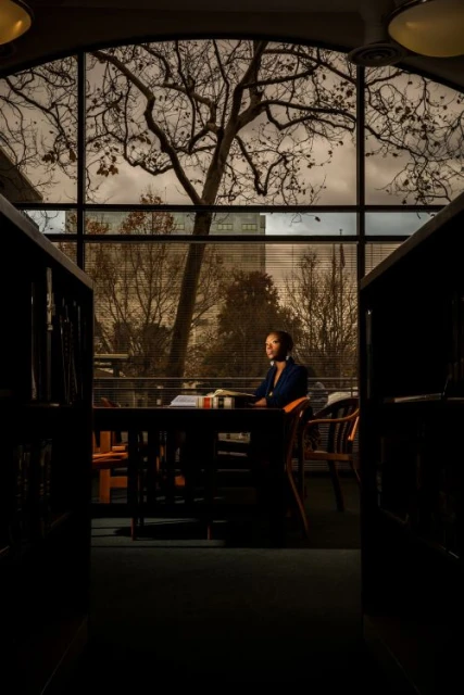 Adult woman sits at a table near a window in a library; bare trees and a cityscape visible through the window in natural, soft lighting.