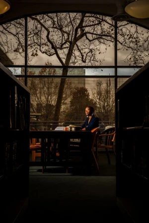 Adult woman sits at a table near a window in a library; bare trees and a cityscape visible through the window in natural, soft lighting.