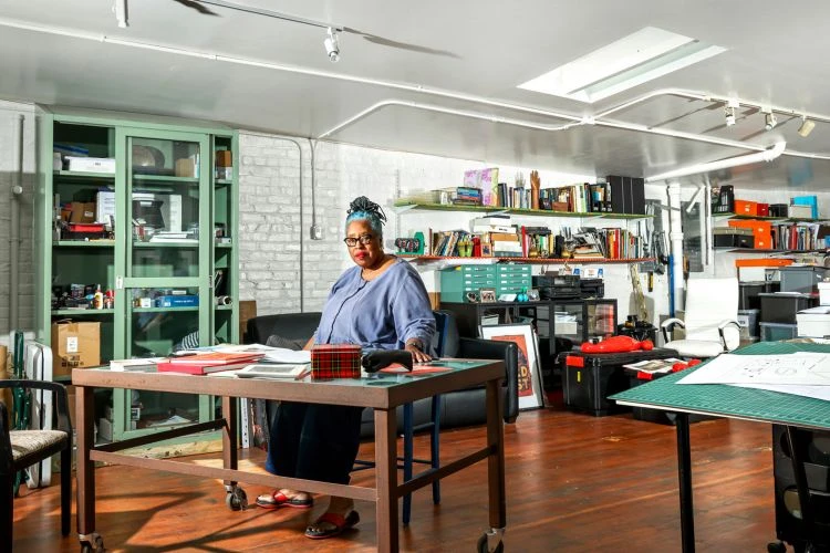Indoor portrait of an adult woman with blue hair in an artist studio. Shelves, artwork, and supplies fill the background of the creative space.