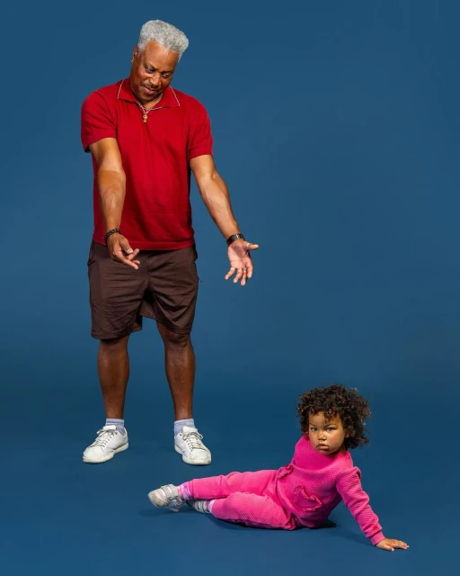 A grandfather in a red shirt gestures downwards at his granddaughter wearing a pink outfit, who is sitting on the blue floor in a studio.