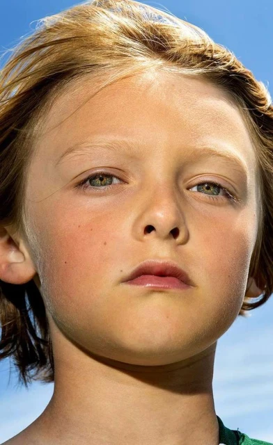 Close-up portrait of a young child with blond hair and fair skin. The kid has a slight frown and is against a blue sky backdrop.