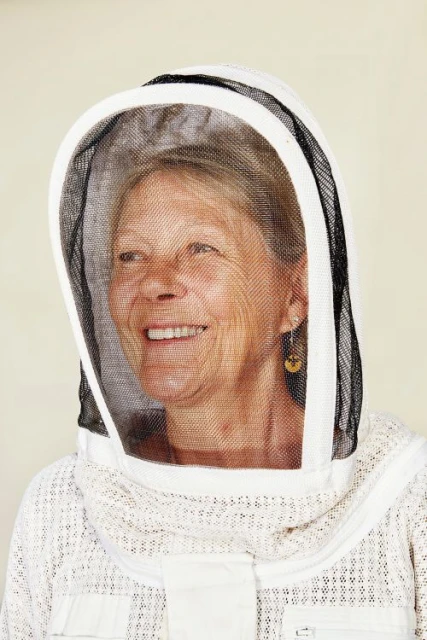Beekeeper with protective mesh hood smiles in close-up portrait against a neutral background.  She wears a white beekeeping suit.