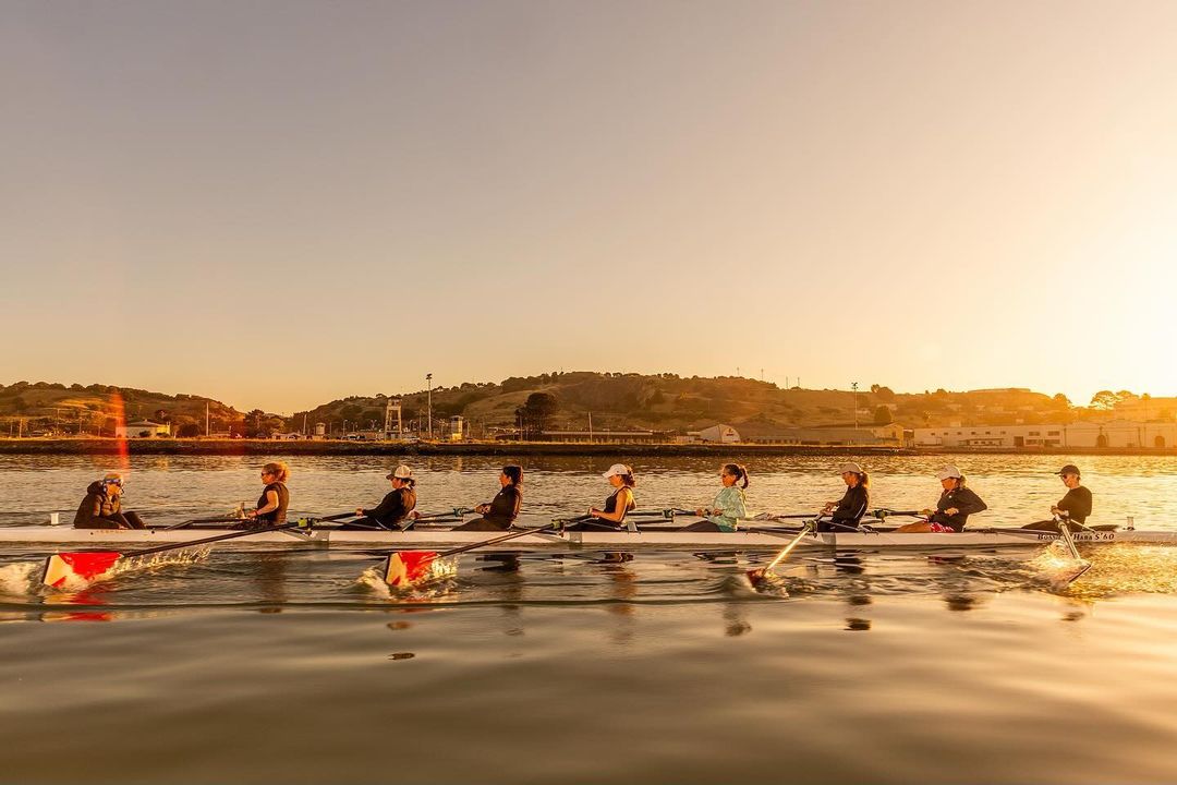 Eight rowers practice on calm water at sunset with a backdrop of trees and buildings in the distance, bathed in golden sunlight.