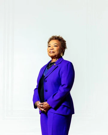 Portrait of a senior African-American woman in a purple suit, standing indoors against a white wall, looking off-camera with hands clasped.