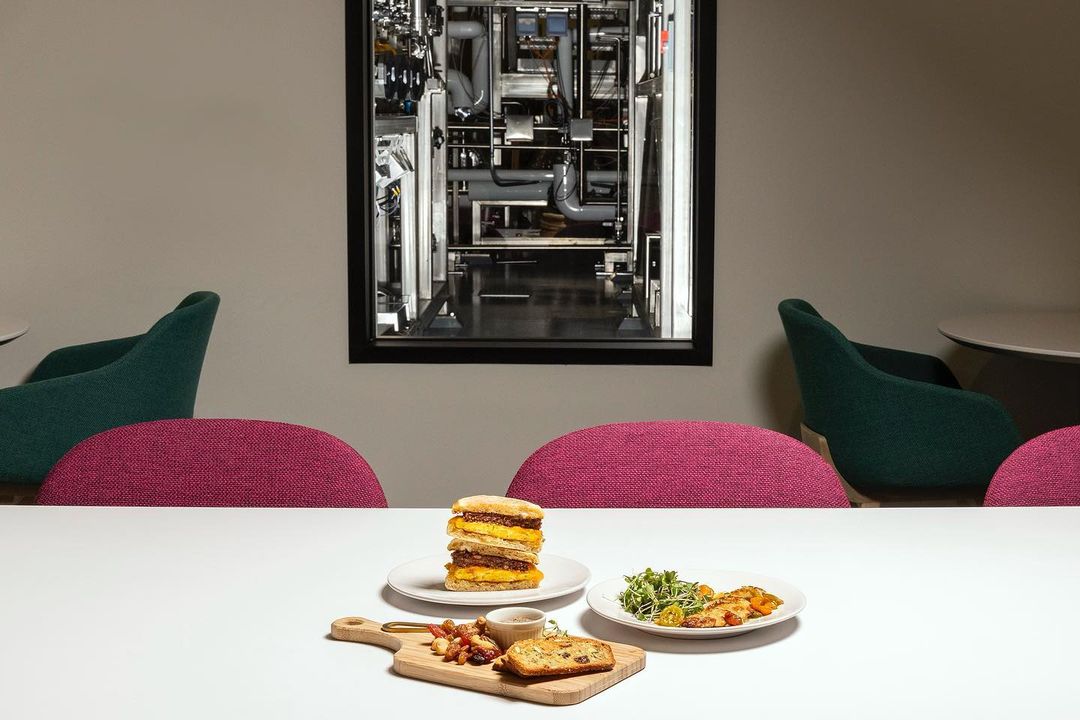 Stacked sandwich and salad on a white table with modern green and pink chairs, beneath a framed monochrome image of industrial pipes.