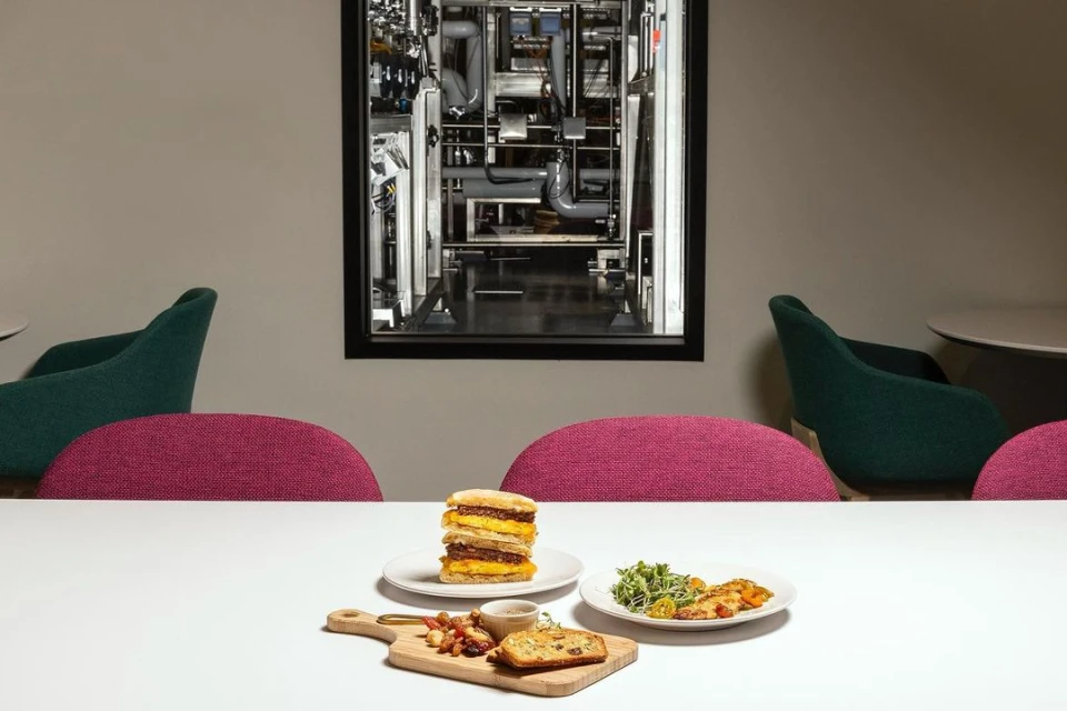 Stacked sandwich and salad on a white table with modern green and pink chairs, beneath a framed monochrome image of industrial pipes.