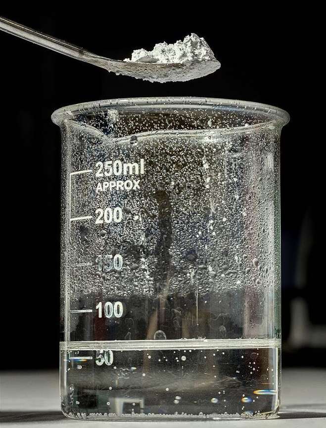 Close-up of a spoon holding a white crystalline substance above a beaker filled with a clear liquid, set against a dark background.
