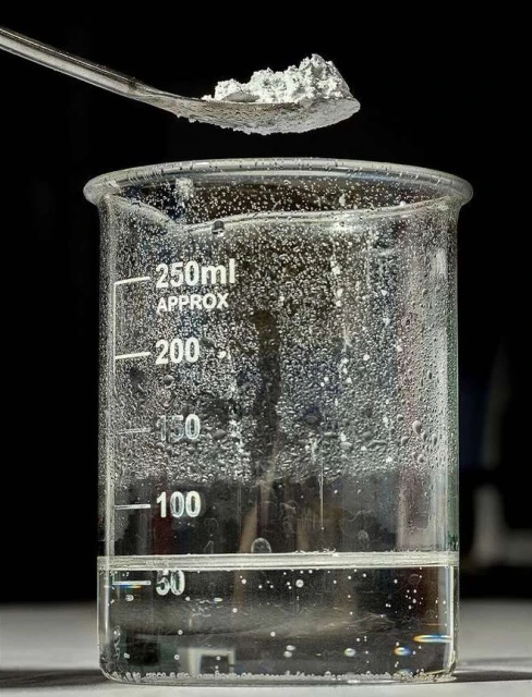 Close-up of a spoon holding a white crystalline substance above a beaker filled with a clear liquid, set against a dark background.