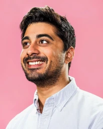 Portrait of a smiling South Asian man wearing a light blue buttoned shirt against a solid pink background.