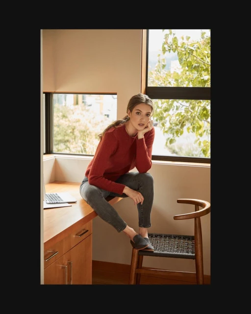 A woman in a red sweater sits on a desk next to a chair, looking at the camera with window views of trees in the background.