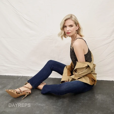Blonde woman seated on dark floor against white fabric backdrop wearing dark jeans, patterned top, and leopard print heels