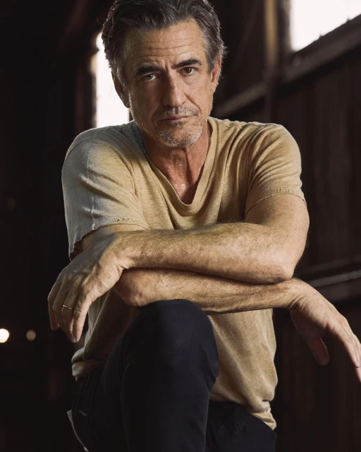 Portrait of a handsome middle-aged man with grey hair, wearing a beige t-shirt, arms crossed, looking at the camera.