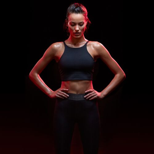 Athletic woman in fitness wear poses with hands on hips in studio with red lighting, dark background. Fitness, sportswear, portrait.