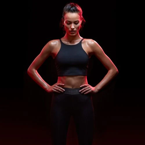 Athletic woman in fitness wear poses with hands on hips in studio with red lighting, dark background. Fitness, sportswear, portrait.