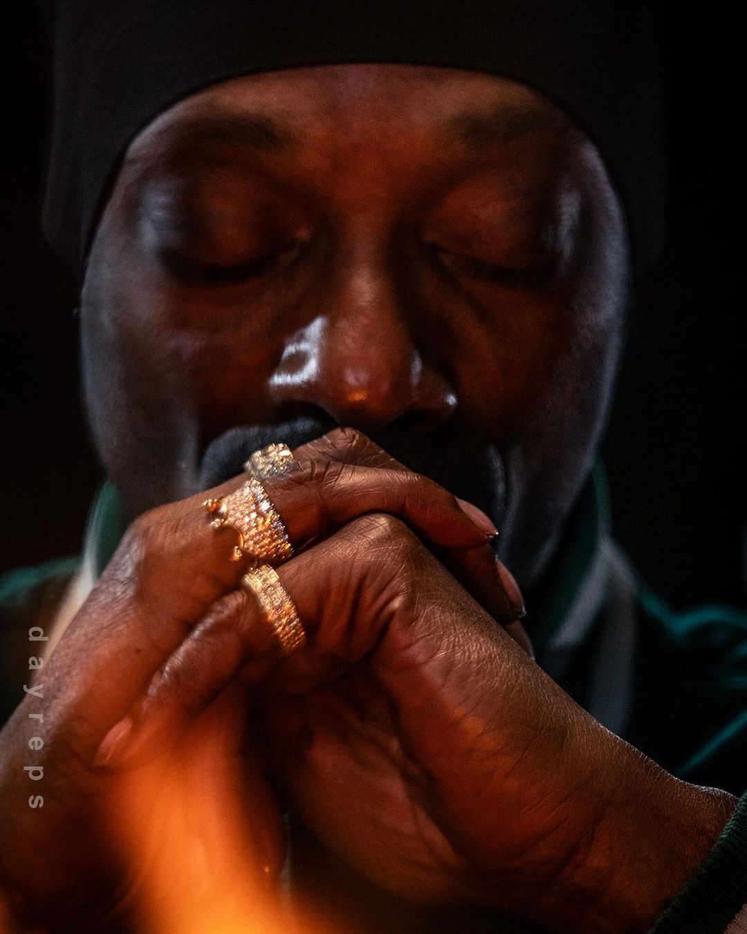 Close-up portrait of an adult male with closed eyes wearing rings on his interlocked hands. Shot with indoor lighting and a dark background.