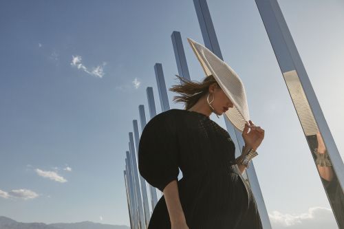A woman wearing a large white hat and dark dress poses outdoors with modern silver pillars against a sunny blue sky backdrop.