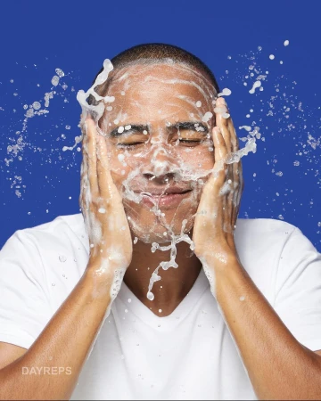 Close-up shot of a smiling man in a white t-shirt washing his face with water and soap on a blue background, showcasing skincare.