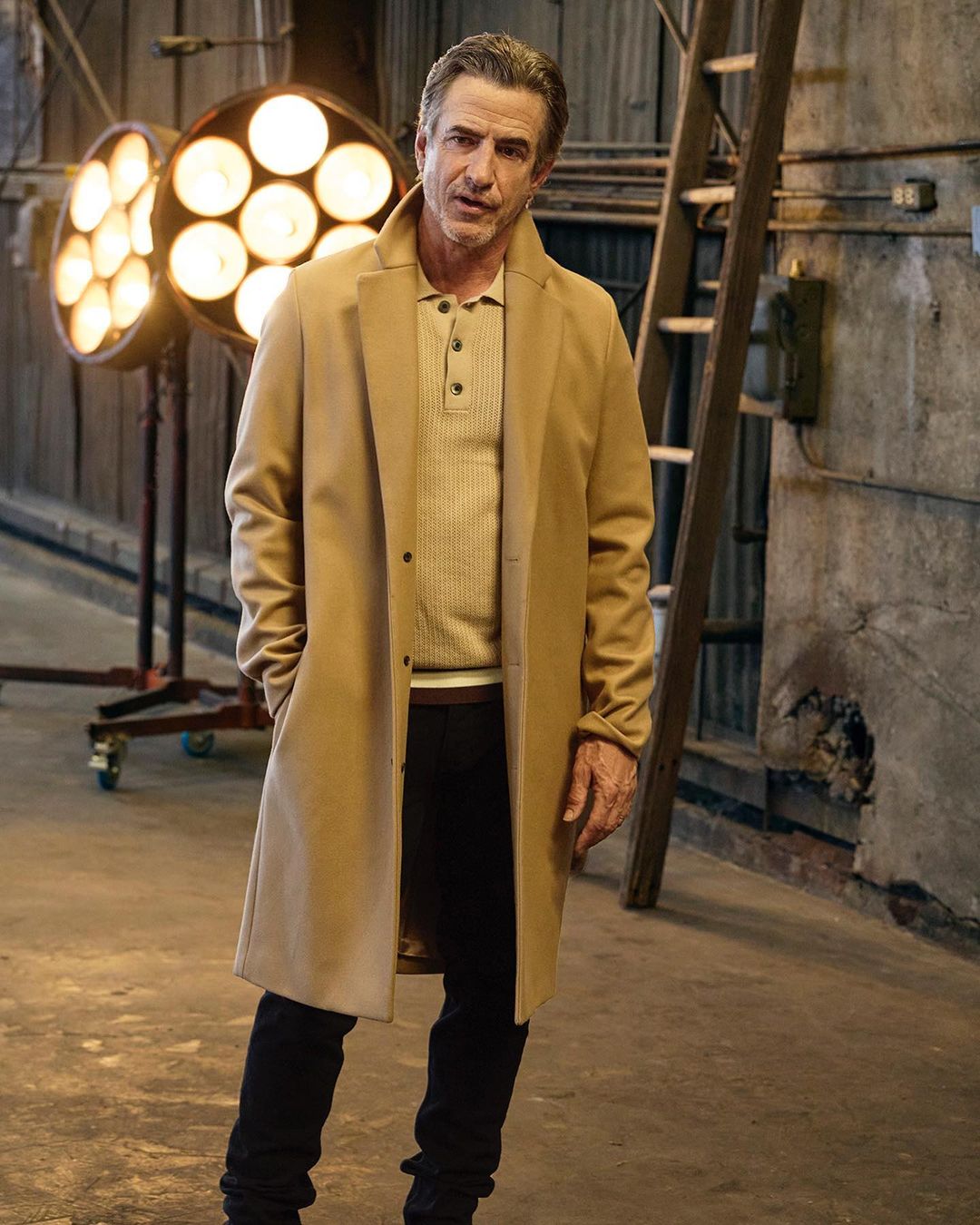 A man in a tan overcoat, light brown ribbed sweater, and dark pants stands indoors near studio lighting, posing for a fashion portrait.