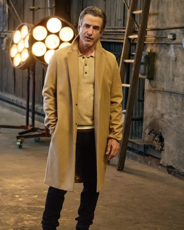 A man in a tan overcoat, light brown ribbed sweater, and dark pants stands indoors near studio lighting, posing for a fashion portrait.