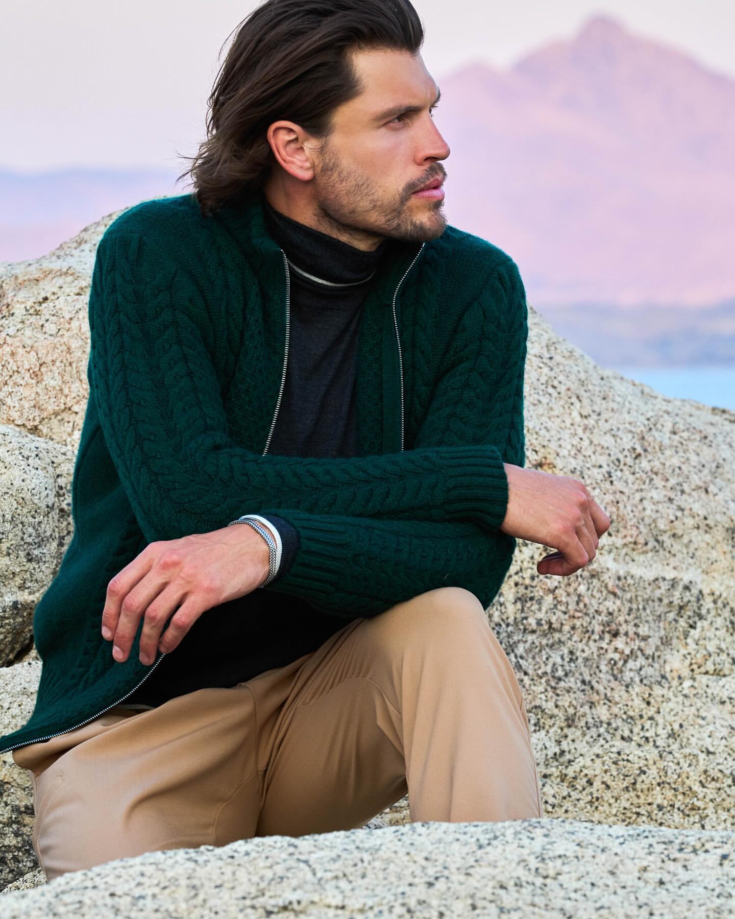 An adult male fashion model poses outdoors, wearing a dark green knit zip-up cardigan, sitting on rocks with mountains visible in background.