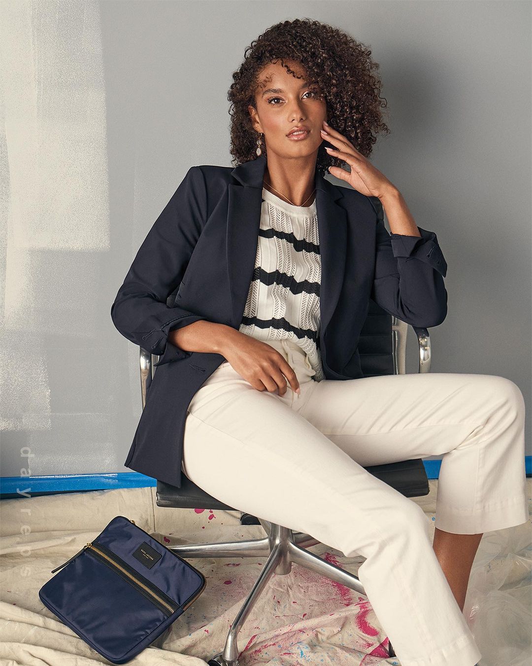 A curly-haired woman in a blazer and white pants poses in a studio, seated on an office chair next to a navy laptop bag.