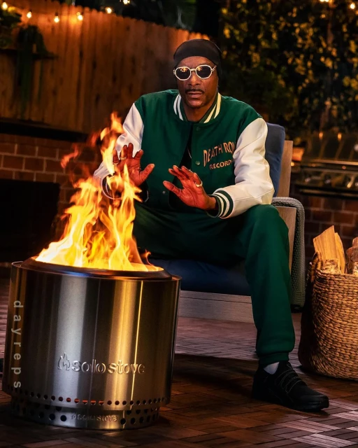 Man sitting by a Solo Stove fire pit in outdoor setting, wearing green jacket and pants, illuminated by warm, inviting light.