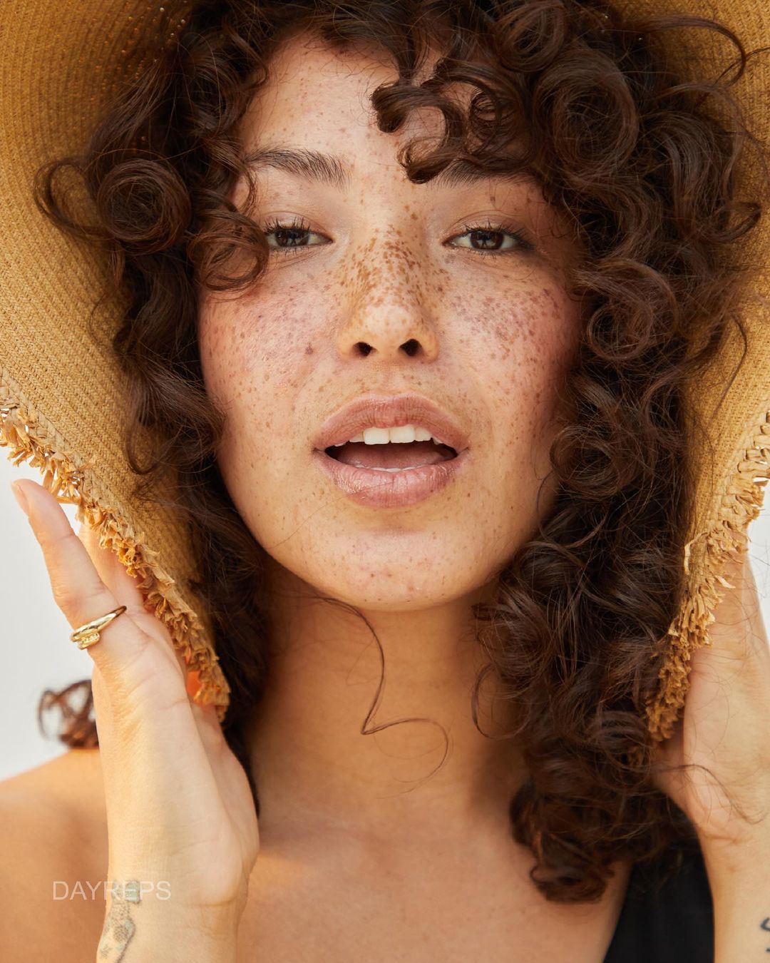 Close-up portrait of a freckled woman with curly brown hair wearing a tan straw hat, against a bright, neutral background.