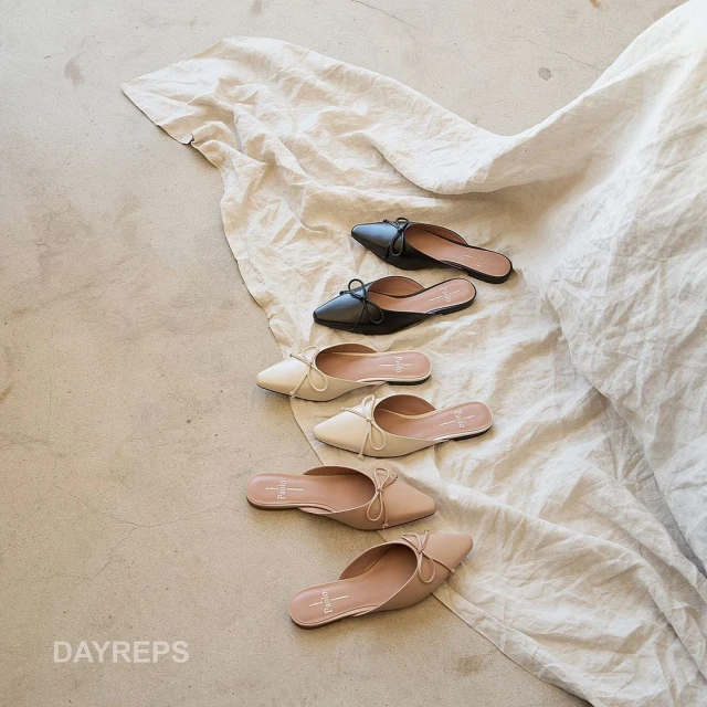 Assortment of stylish women's mule shoes with bows in black, beige, and off-white arranged on a neutral wrinkled fabric backdrop, product shot.