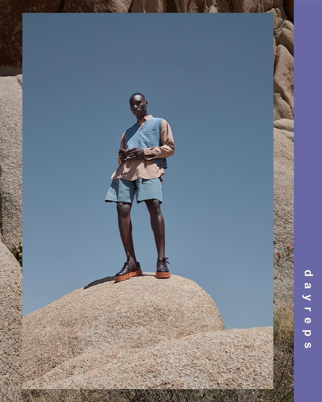 An adult African-American male stands on a desert boulder. He wears light-colored shorts, vest, and shirt against a clear blue sky.