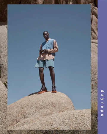 An adult African-American male stands on a desert boulder. He wears light-colored shorts, vest, and shirt against a clear blue sky.