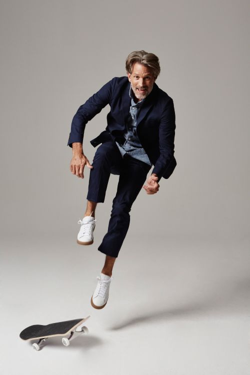 Adult male in a navy suit and white sneakers jumping over a skateboard in a light grey studio setting, showcasing action and fashion.