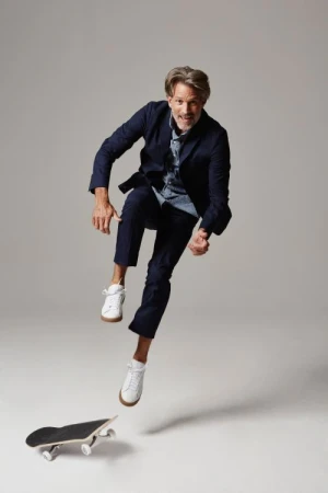 Adult male in a navy suit and white sneakers jumping over a skateboard in a light grey studio setting, showcasing action and fashion.