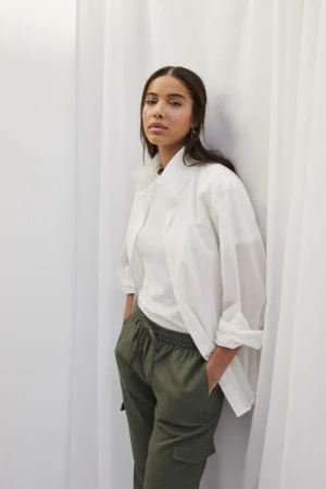 A woman poses in a white collared shirt and olive green cargo pants against a neutral background, offering a relaxed yet stylish look.