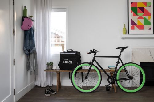 Uber Eats delivery bag and black bicycle with green tires indoors near a bench, wall art, shoes, and a white wall.