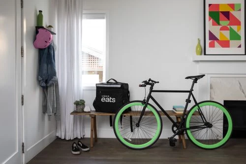 Uber Eats delivery bag and black bicycle with green tires indoors near a bench, wall art, shoes, and a white wall.